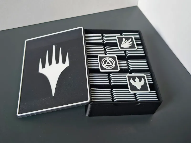 Schwarz-weiß gedruckte Box für Spielmarker: links der Deckel mit weißem Symbol, rechts das offene Fach voller gestapelter Plättchen
